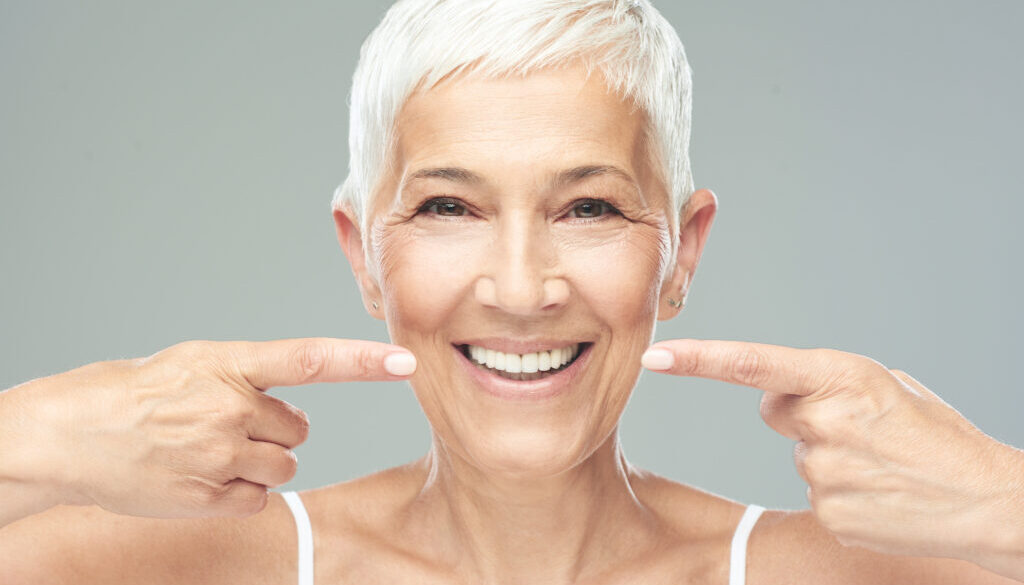 Beautiful Caucasian smiling senior woman with short grey hair pointing at her teeth and looking at camera. Beauty photography.